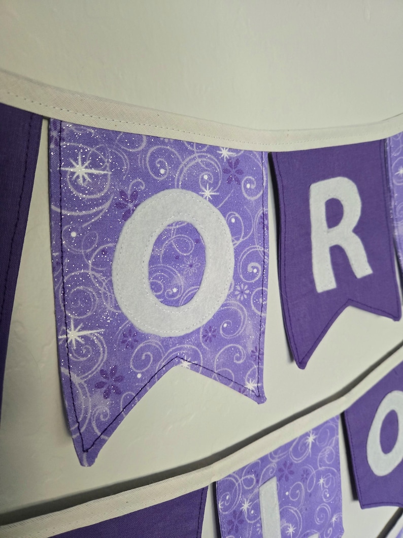 K&ouml;nnte beinhalten: Lila Banner mit wei&szlig;en Buchstaben, die ein Wort bilden. Das Banner hat einen wei&szlig;en Rand und h&auml;ngt an einer wei&szlig;en Wand. Der Buchstabe "O" befindet sich auf einem Banner mit einem Wirbelmuster und der Buchstabe "R" auf einem einfarbigen lila Banner.