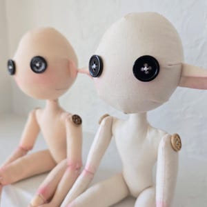 May include: Two handmade fabric dolls with button eyes and button joints. One doll is light pink with blush and the other is off-white. Both dolls have small, pointed ears and are seated.