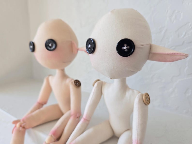 May include: Two handmade fabric dolls with button eyes and button joints. One doll is light pink with blush and the other is off-white. Both dolls have small, pointed ears and are seated.