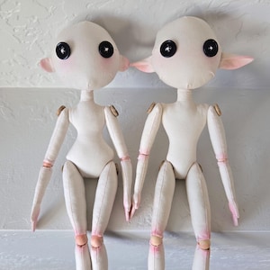 May include: Two handmade cloth dolls with button eyes, pink cheeks, and small, pointed ears. The dolls have articulated limbs with wooden joints and pink accents on the hands and legs. They stand side-by-side against a neutral background.
