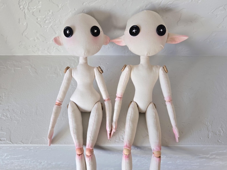 May include: Two handmade cloth dolls with button eyes, pink cheeks, and small, pointed ears. The dolls have articulated limbs with wooden joints and pink accents on the hands and legs. They stand side-by-side against a neutral background.