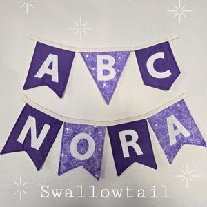 K&ouml;nnte beinhalten: Lila Stoffbanner mit den wei&szlig;en Buchstaben "ABC" &uuml;ber "NORA". Das mittlere Banner hat ein wei&szlig;es Blumenmuster. Das Banner ist an einem wei&szlig;en Stoff befestigt. Unter dem Banner steht das Wort "Swallowtail".