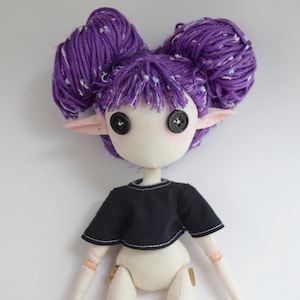 May include: A handcrafted doll with pale skin, pointed ears, and two large purple yarn buns. The doll features black button eyes and wears a short-sleeved black crop top with visible white stitching along the hem and neckline.
