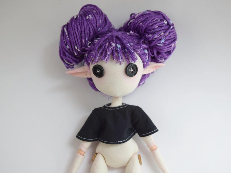 May include: A handcrafted doll with pale skin, pointed ears, and two large purple yarn buns. The doll features black button eyes and wears a short-sleeved black crop top with visible white stitching along the hem and neckline.