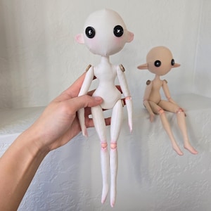 May include: Two handmade dolls with button eyes and articulated limbs. One doll is held by a hand, the other is seated. The dolls have a minimalist design, with a white body and pink accents on the ears and joints. The seated doll is tan.