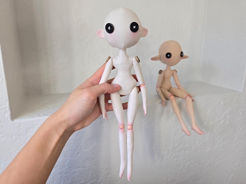 May include: Two handmade dolls with button eyes and articulated limbs. One doll is held by a hand, the other is seated. The dolls have a minimalist design, with a white body and pink accents on the ears and joints. The seated doll is tan.