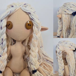 May include: A handmade cloth doll with long, braided, cream-colored hair with purple flecks. The doll has large button eyes, pointed ears, and a simple, neutral-toned body. Dark blue ribbons accent the braids.