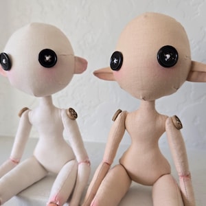 May include: Two handmade fabric dolls with button eyes and button joints. One doll is off-white, the other is tan. Both have pink cheeks and ears. The dolls are sitting side by side.