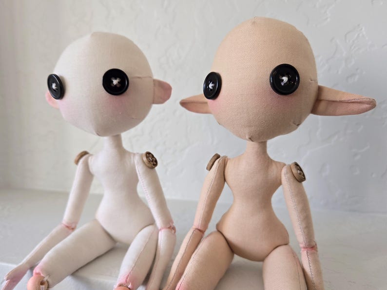 May include: Two handmade fabric dolls with button eyes and button joints. One doll is off-white, the other is tan. Both have pink cheeks and ears. The dolls are sitting side by side.