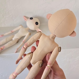 May include: Two unfinished cloth dolls with articulated limbs and button joints. One doll is held in a hand, showcasing the tan fabric body and head with small ears. The other doll is in the background, with a white body and head.
