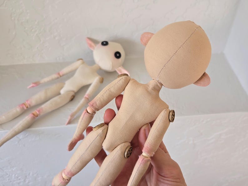 May include: Two unfinished cloth dolls with articulated limbs and button joints. One doll is held in a hand, showcasing the tan fabric body and head with small ears. The other doll is in the background, with a white body and head.