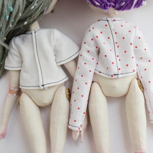 May include: Two fabric dolls viewed from the back. The left doll has sage green yarn hair and wears a plain white short-sleeved top. The right doll has purple yarn hair and wears a white long-sleeved top with a small red polka dot pattern.