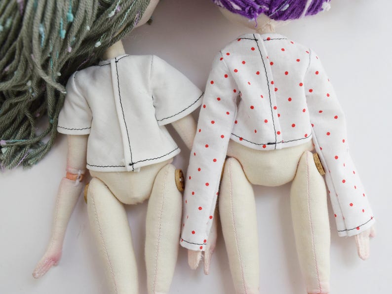 May include: Two fabric dolls viewed from the back. The left doll has sage green yarn hair and wears a plain white short-sleeved top. The right doll has purple yarn hair and wears a white long-sleeved top with a small red polka dot pattern.