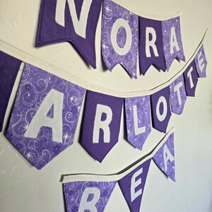 K&ouml;nnte beinhalten: Lila Stoffbanner mit wei&szlig;en Buchstaben, die "NORA", "CHARLOTTE" und "BEA" buchstabieren. Die Bannerfahnen sind lila mit wei&szlig;en Buchstaben, und einige Fahnen haben ein gemustertes Design. Das Banner h&auml;ngt an einer wei&szlig;en Wand.