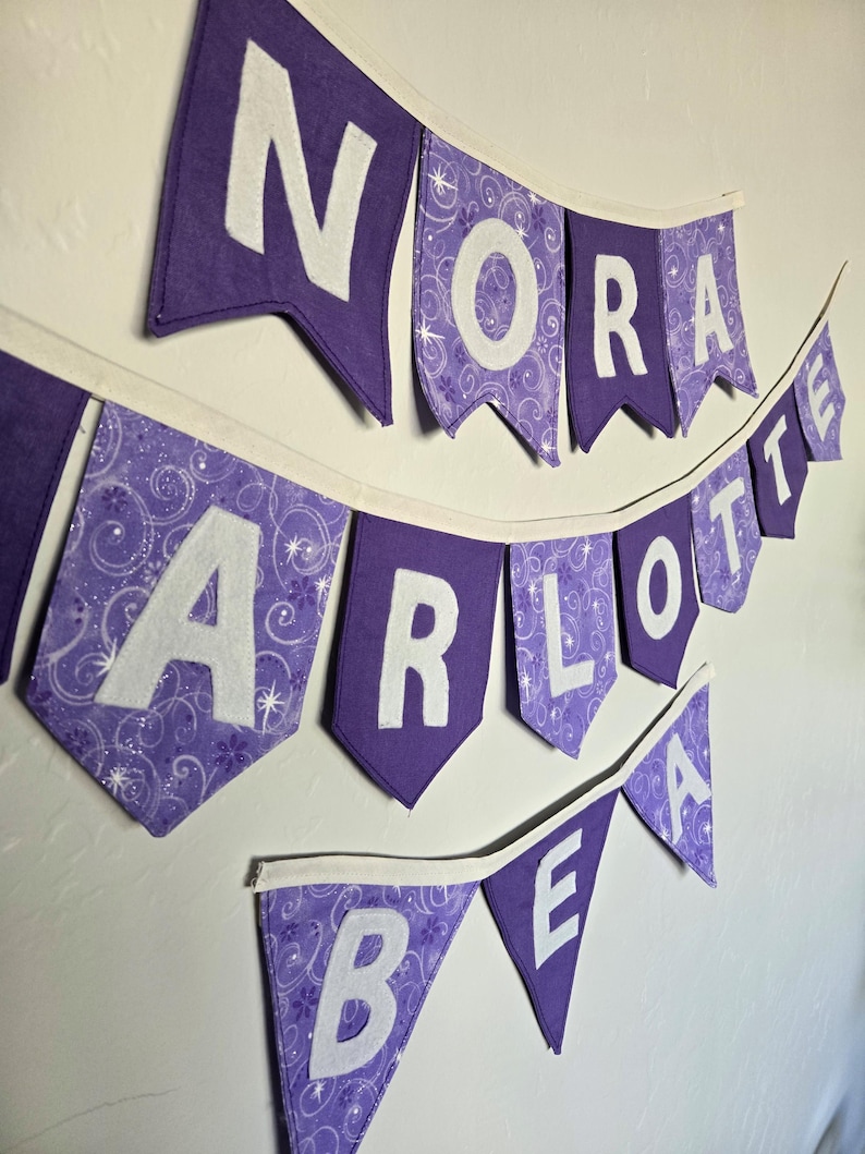 K&ouml;nnte beinhalten: Lila Stoffbanner mit wei&szlig;en Buchstaben, die "NORA", "CHARLOTTE" und "BEA" buchstabieren. Die Bannerfahnen sind lila mit wei&szlig;en Buchstaben, und einige Fahnen haben ein gemustertes Design. Das Banner h&auml;ngt an einer wei&szlig;en Wand.