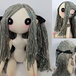 May include: A handmade cloth doll with a whimsical design. The doll has a pale complexion, large button eyes, and gray yarn hair with colorful flecks. Black bows adorn the hair. The doll's body is a light beige color.