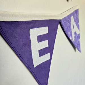 K&ouml;nnte beinhalten: Lila Stoffbanner mit wei&szlig;en Buchstaben, die "E A" buchstabieren. Das Banner ist dreieckig, mit einem wei&szlig;en Rand entlang der Oberkante. Die Buchstaben bestehen aus wei&szlig;em Filz und sind auf den lila Stoff gen&auml;ht.