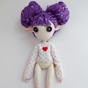 May include: A handmade doll with purple yarn hair styled in two buns, black button eyes, and pointed ears. The doll wears a white long-sleeved shirt featuring a pattern of small red dots and a single red heart appliqu&eacute; centered on the chest area.