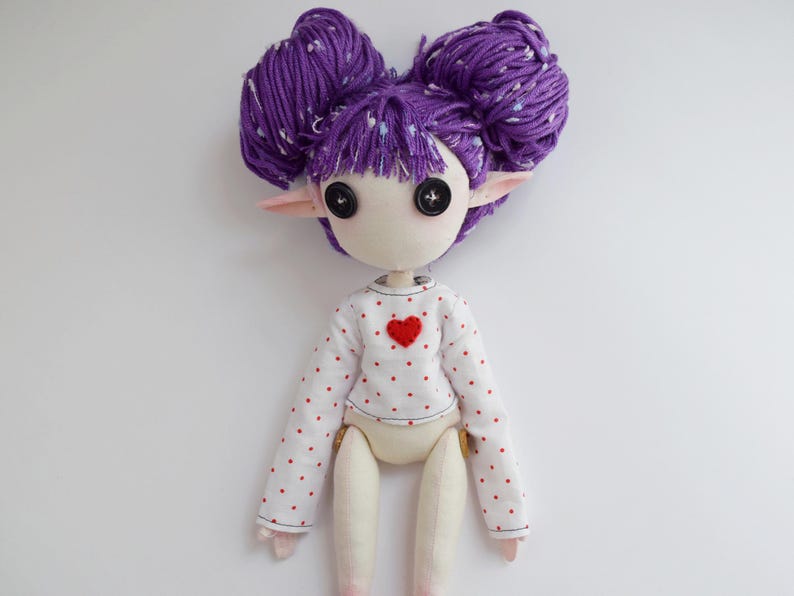 May include: A handmade doll with purple yarn hair styled in two buns, black button eyes, and pointed ears. The doll wears a white long-sleeved shirt featuring a pattern of small red dots and a single red heart appliqu&eacute; centered on the chest area.