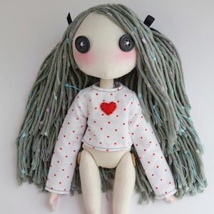 May include: A handmade fabric doll with long, textured sage green yarn hair featuring small blue and purple accents. The doll has black button eyes, pink cheeks, and wears a white long-sleeve shirt with a red polka dot pattern and a red heart.