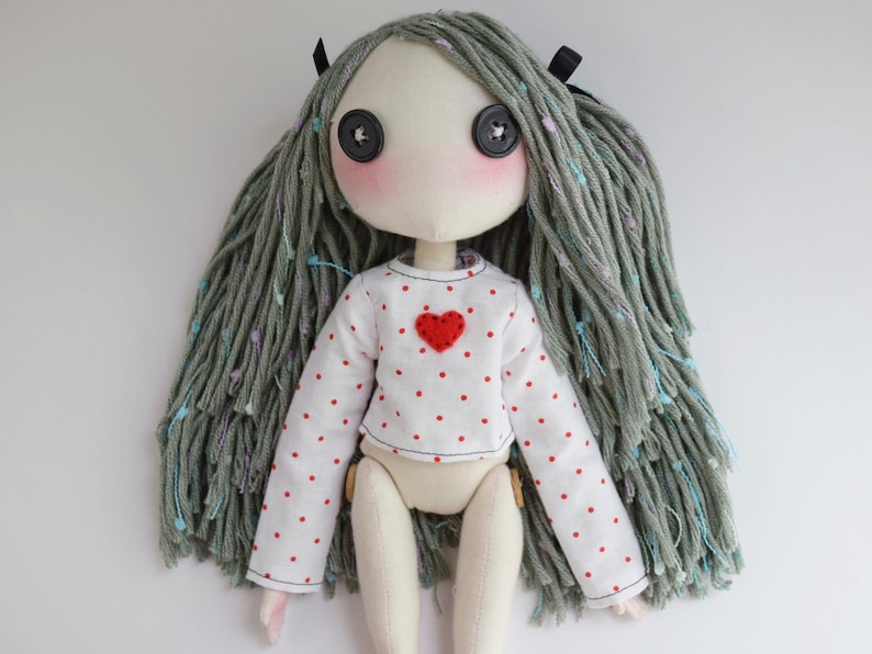 May include: A handmade fabric doll with long, textured sage green yarn hair featuring small blue and purple accents. The doll has black button eyes, pink cheeks, and wears a white long-sleeve shirt with a red polka dot pattern and a red heart.