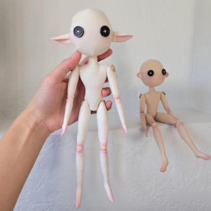 May include: Two handmade art dolls. One doll is held up, featuring a white body, large black button eyes, and pointed ears. The other doll is seated, with a tan body and similar black button eyes. Both dolls have articulated joints.