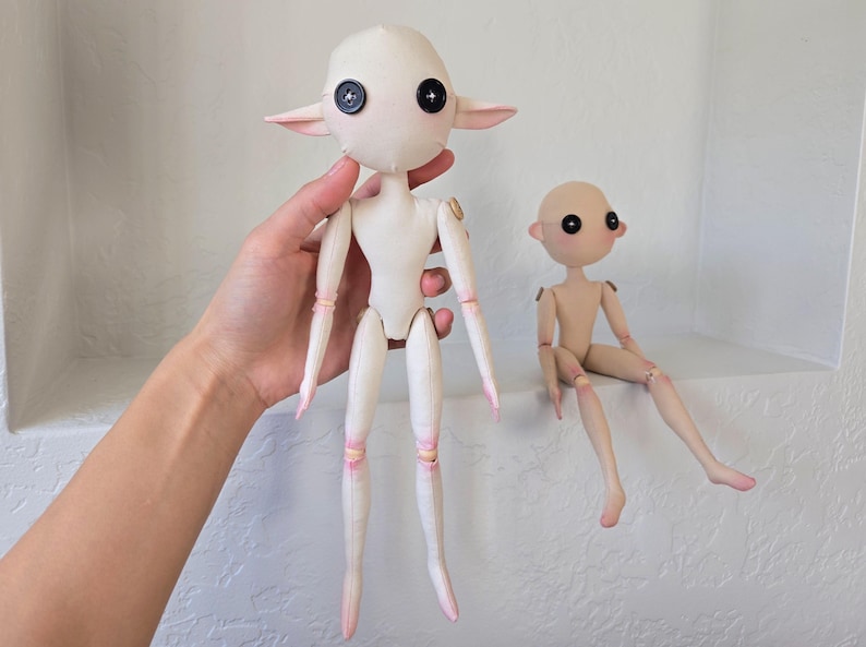 May include: Two handmade art dolls. One doll is held up, featuring a white body, large black button eyes, and pointed ears. The other doll is seated, with a tan body and similar black button eyes. Both dolls have articulated joints.
