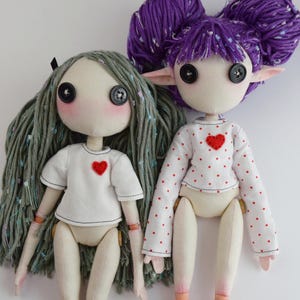 May include: Two handmade dolls with button eyes and yarn hair. The left doll has long sage green hair and a white t-shirt with a red heart. The right doll has purple hair styled in buns, pointed ears, and a white long-sleeve shirt with red polka dots and a red heart.