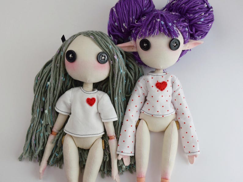 May include: Two handmade dolls with button eyes and yarn hair. The left doll has long sage green hair and a white t-shirt with a red heart. The right doll has purple hair styled in buns, pointed ears, and a white long-sleeve shirt with red polka dots and a red heart.