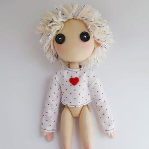 May include: A handmade fabric doll with a beige complexion, black button eyes, and messy cream-colored yarn hair. The doll wears a white long-sleeved crop top featuring a pattern of small red dots and a single red heart appliqu&eacute; on the chest area.