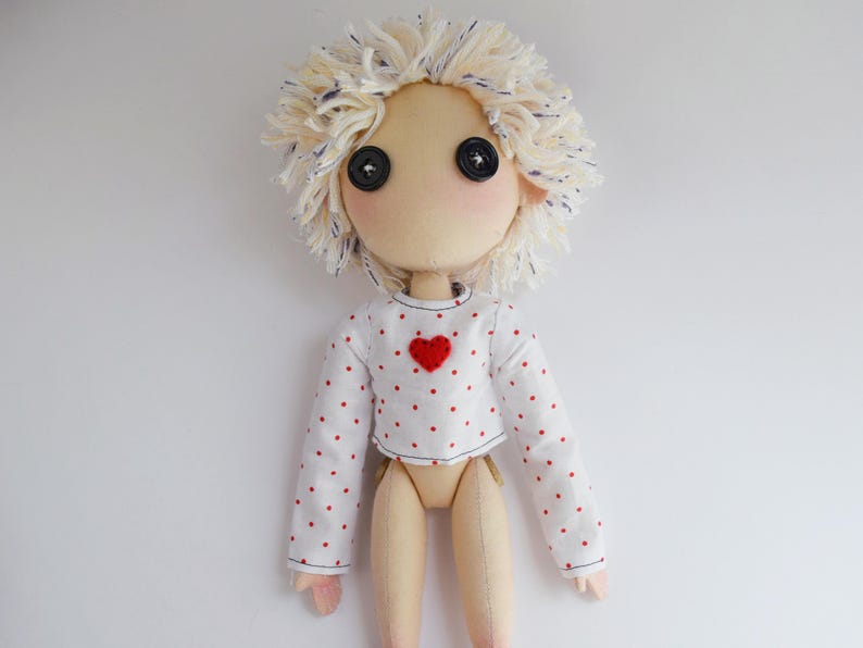 May include: A handmade fabric doll with a beige complexion, black button eyes, and messy cream-colored yarn hair. The doll wears a white long-sleeved crop top featuring a pattern of small red dots and a single red heart appliqu&eacute; on the chest area.