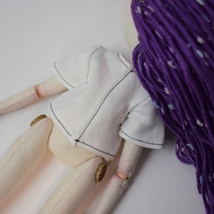 May include: A fabric doll with light beige skin, wearing a short-sleeved white shirt with visible black stitching. The doll features long, textured purple yarn hair with white flecks and small round wooden buttons at the hip joints.
