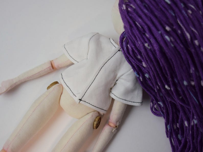 May include: A fabric doll with light beige skin, wearing a short-sleeved white shirt with visible black stitching. The doll features long, textured purple yarn hair with white flecks and small round wooden buttons at the hip joints.