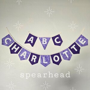 K&ouml;nnte beinhalten: Lila-wei&szlig;es Stoffbanner mit den Buchstaben "CHARLOTTE" und "ABC" mit einem dekorativen Muster. Das Wort "spearhead" steht darunter. Wei&szlig;e Sternausbr&uuml;che sind um das Banner herum verstreut.