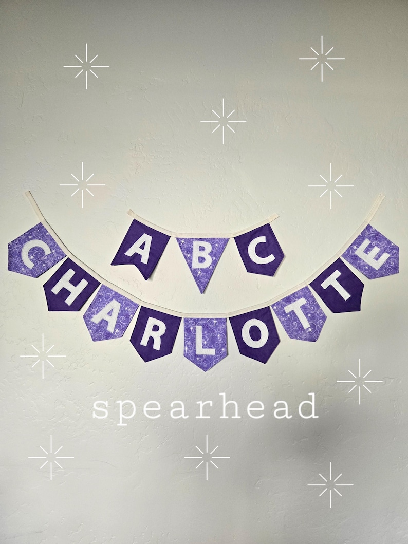 K&ouml;nnte beinhalten: Lila-wei&szlig;es Stoffbanner mit den Buchstaben "CHARLOTTE" und "ABC" mit einem dekorativen Muster. Das Wort "spearhead" steht darunter. Wei&szlig;e Sternausbr&uuml;che sind um das Banner herum verstreut.