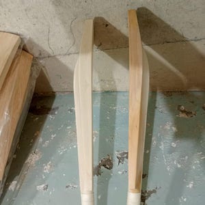 May include: Two wooden cricket bats, one light-colored and the other with a darker, natural wood tone. Both bats have white grip tape on the handles. The bats are leaning against a textured, light-colored wall.