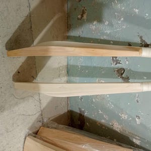 May include: Two wooden cricket bats with light-colored wood blades and wrapped handles are displayed against a textured, light blue-green wall. The bats are positioned horizontally, showcasing their curved design and natural wood grain.