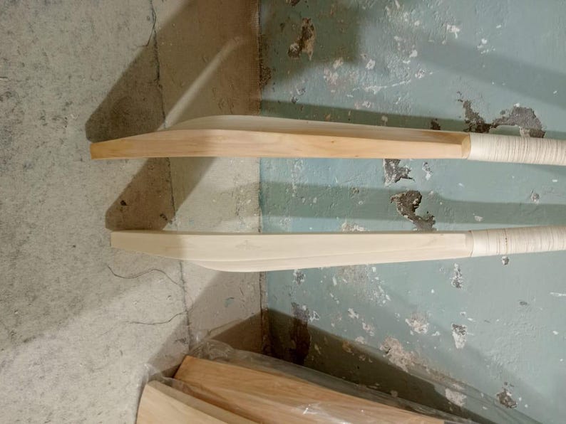 May include: Two wooden cricket bats with light-colored wood blades and wrapped handles are displayed against a textured, light blue-green wall. The bats are positioned horizontally, showcasing their curved design and natural wood grain.