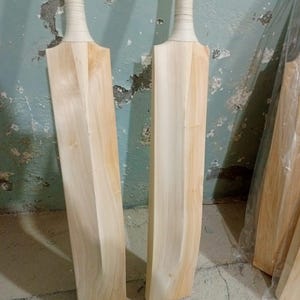 May include: Two unfinished cricket bats made of light-coloured wood. The handles are wrapped in a light-coloured grip. The bats are standing upright against a textured, light blue wall.