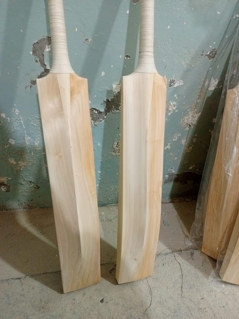 May include: Two unfinished cricket bats made of light-colored wood. The handles are wrapped in a light-colored grip. The bats are standing upright against a textured, light blue wall.