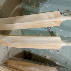 May include: Two unfinished wooden cricket bats with light brown blades and wrapped handles. The bats are positioned at an angle, showcasing their shape and design. A stack of additional bats is visible in the background.