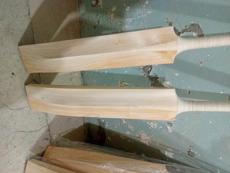 May include: Two unfinished wooden cricket bats with light brown blades and wrapped handles. The bats are positioned at an angle, showcasing their shape and design. A stack of additional bats is visible in the background.