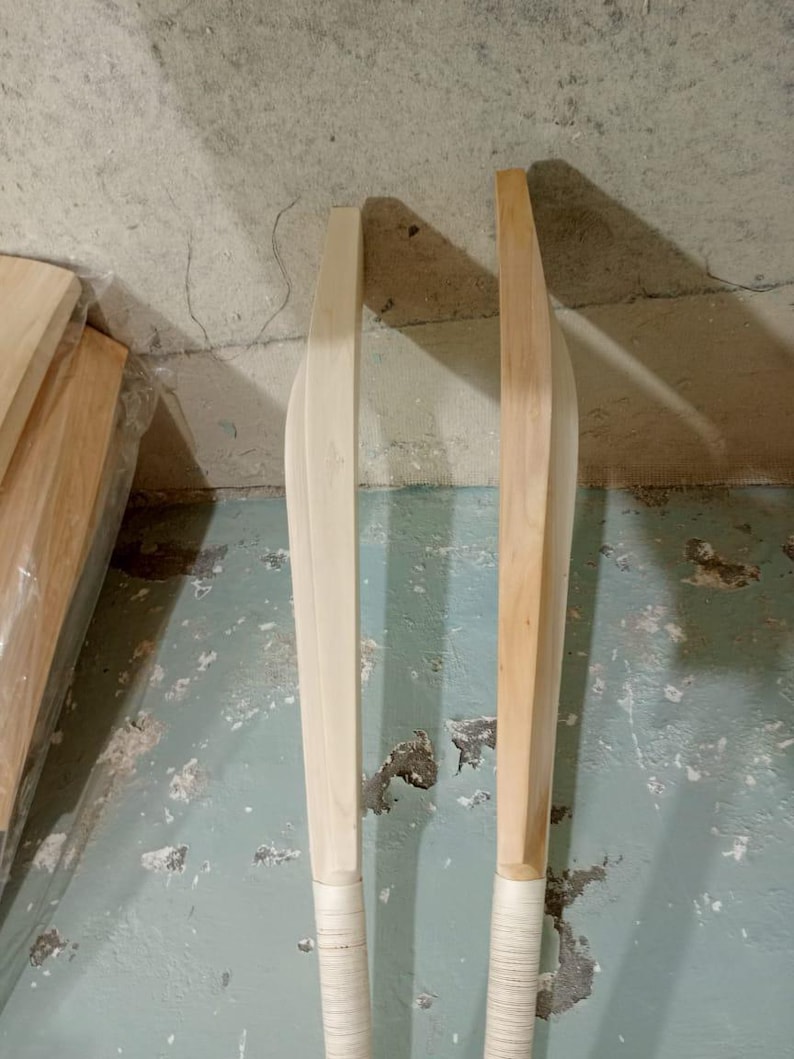 May include: Two wooden cricket bats, one light-colored and the other with a darker, natural wood tone. Both bats have a rounded blade and a wrapped grip. The bats are leaning against a textured, light-colored wall.