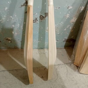 May include: Two wooden cricket bats with white grips. One bat is a light brown color, and the other is a natural wood color. The bats are standing upright against a textured, light blue wall.
