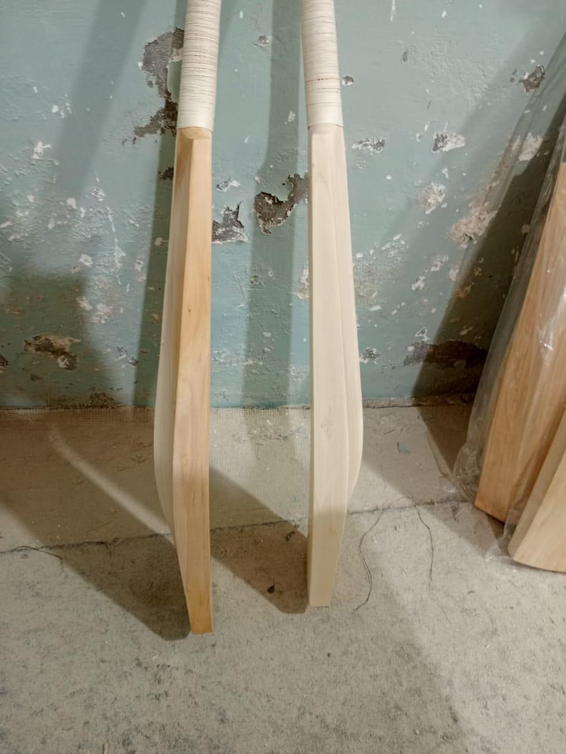 May include: Two wooden cricket bats with white grips. One bat is a light brown color, and the other is a natural wood color. The bats are standing upright against a textured, light blue wall.