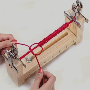 May include: A wooden macrame board with a red cord secured by metal clips. The board has a groove for the cord and measurement markings. A section of the cord is braided, and the other end is being worked on.