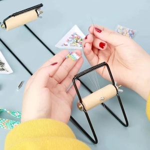 May include: A bead loom with a wooden roller and black metal frame is shown. The image also shows a person's hands holding a needle and beads. The beads are in various colors, and the loom is used for crafting beaded jewelry.