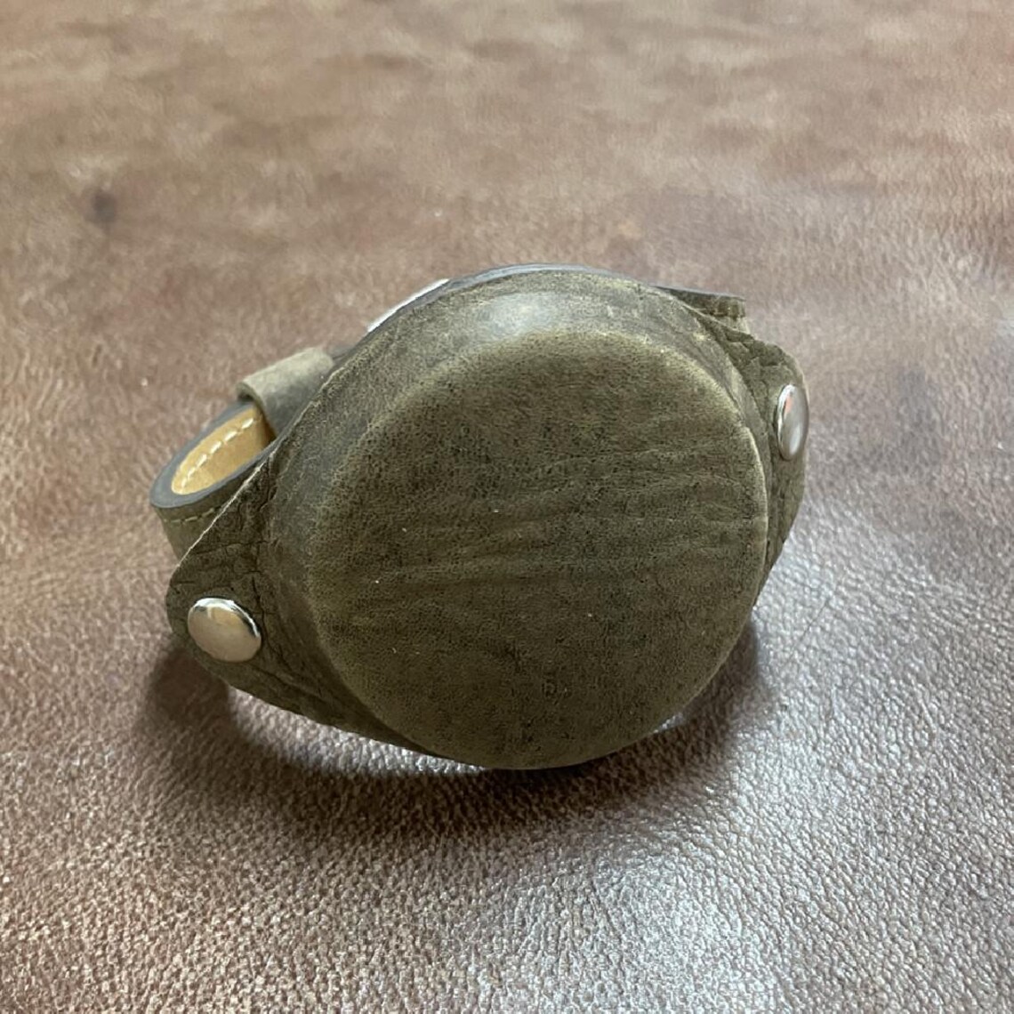 May include: A dark green leather watch band with silver metal accents. The band is wrapped around a round, dark green leather watch face.