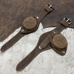 Puede incluir: Dos correas de reloj de cuero marrón con cubiertas circulares integradas. Cada correa presenta un diseño de brazalete ancho, hebillas de metal plateado y remaches plateados que aseguran los protectores de esfera a las bandas de cuero.