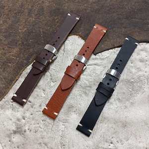 May include: Three leather watch straps in brown, tan, and black. Each strap features a silver buckle and white stitching along the edges. The straps are displayed on a textured, brown surface.